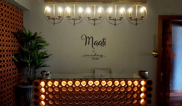 A warmly lit reception area with a modern, minimalist design. An illuminated circular chandelier hangs above a front desk with a decorative lattice pattern. A plant in the corner adds a touch of greenery beside brick-style wall accents. The words 'Maati by sundays the spa' are displayed on the wall.