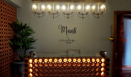 A warmly lit reception area with a modern, minimalist design. An illuminated circular chandelier hangs above a front desk with a decorative lattice pattern. A plant in the corner adds a touch of greenery beside brick-style wall accents. The words 'Maati by sundays the spa' are displayed on the wall.