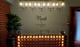A warmly lit reception area with a modern, minimalist design. An illuminated circular chandelier hangs above a front desk with a decorative lattice pattern. A plant in the corner adds a touch of greenery beside brick-style wall accents. The words 'Maati by sundays the spa' are displayed on the wall.