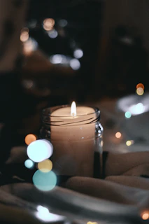 A soft-lit candle in a glass jar resting on a light wooden table with subtle shadows.
