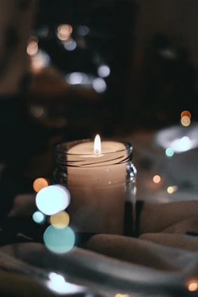 A soft-lit candle in a glass jar resting on a light wooden table with subtle shadows.