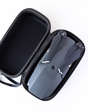 A compact drone with folded arms is neatly placed inside an open black carrying case with a zipper. The drone is sleek and modern, featuring a dark gray color with minor detailing on its surface.