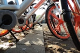 A row of rental bicycles with silver frames and bright orange wheels and accents are parked on the side of a street. The ground is covered with dry autumn leaves, casting shadows from the sunlight. In the background, a few people are walking past, suggesting an urban setting.