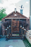 A rustic wooden structure resembling a small house or shed with a prominent wooden door at the center. Its design is complemented by a weathered skull of a ram or similar animal mounted at the roof's peak. There are twig brooms with handwritten signs attached along the entrance pathway on each side. A unique figure resembling a witch or folklore character is visible on top of the chimney, adding a whimsical touch to the setting. The setting is outdoors with a partially cloudy sky.
