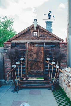 A rustic wooden structure resembling a small house or shed with a prominent wooden door at the center. Its design is complemented by a weathered skull of a ram or similar animal mounted at the roof's peak. There are twig brooms with handwritten signs attached along the entrance pathway on each side. A unique figure resembling a witch or folklore character is visible on top of the chimney, adding a whimsical touch to the setting. The setting is outdoors with a partially cloudy sky.
