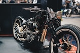 A custom motorcycle with a glossy black finish highlighted by golden accents and intricate detailing. The design features a retro aesthetic, with wide tires and a polished metallic engine. The setting appears to be a showroom or exhibition space with various other motorcycles visible in the background.