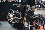A custom motorcycle with a glossy black finish highlighted by golden accents and intricate detailing. The design features a retro aesthetic, with wide tires and a polished metallic engine. The setting appears to be a showroom or exhibition space with various other motorcycles visible in the background.