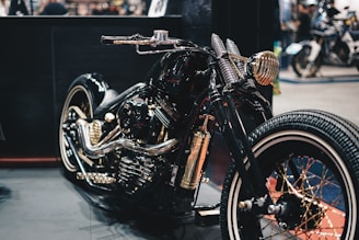 A custom motorcycle with a glossy black finish highlighted by golden accents and intricate detailing. The design features a retro aesthetic, with wide tires and a polished metallic engine. The setting appears to be a showroom or exhibition space with various other motorcycles visible in the background.