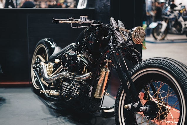 A custom motorcycle with a glossy black finish highlighted by golden accents and intricate detailing. The design features a retro aesthetic, with wide tires and a polished metallic engine. The setting appears to be a showroom or exhibition space with various other motorcycles visible in the background.