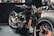 A custom motorcycle with a glossy black finish highlighted by golden accents and intricate detailing. The design features a retro aesthetic, with wide tires and a polished metallic engine. The setting appears to be a showroom or exhibition space with various other motorcycles visible in the background.