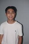 A young person is standing against a plain gray background, wearing a white T-shirt with a small logo on the chest. The person has short dark hair and is smiling, showcasing braces.