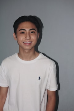 A young person is standing against a plain gray background, wearing a white T-shirt with a small logo on the chest. The person has short dark hair and is smiling, showcasing braces.