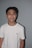 A young person is standing against a plain gray background, wearing a white T-shirt with a small logo on the chest. The person has short dark hair and is smiling, showcasing braces.