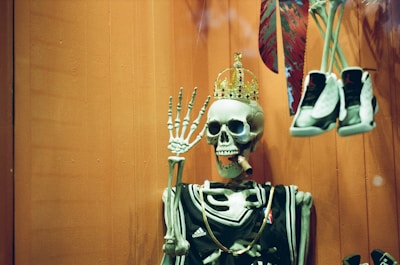 A skeleton wearing a sports jersey and a golden crown with jewels while holding a cigar in its mouth. The skeleton is positioned in front of an orangish backdrop. A pair of sporty shoes are hanging to its right side. The display seems humorous and slightly surreal.