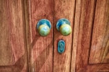 A pair of old, round brass door knobs mounted on a richly-textured wooden door with visible grain patterns. The metal shows signs of tarnishing and weathering, contributing to a rustic and vintage appearance. A small keyhole is situated between the knobs.
