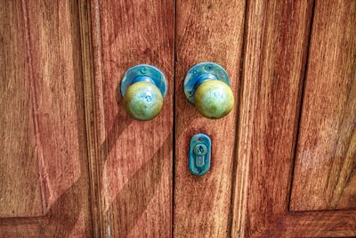 A pair of old, round brass door knobs mounted on a richly-textured wooden door with visible grain patterns. The metal shows signs of tarnishing and weathering, contributing to a rustic and vintage appearance. A small keyhole is situated between the knobs.