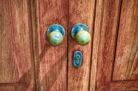 A pair of old, round brass door knobs mounted on a richly-textured wooden door with visible grain patterns. The metal shows signs of tarnishing and weathering, contributing to a rustic and vintage appearance. A small keyhole is situated between the knobs.
