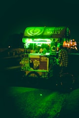 A street-side ice cream cart is illuminated by a bright green light, with a person standing next to it. The cart displays the brand Kwality Wall's and has posters of ice cream products. The background is dark with hints of buildings and people in the distance.