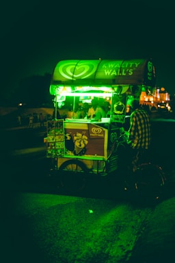 A street-side ice cream cart is illuminated by a bright green light, with a person standing next to it. The cart displays the brand Kwality Wall's and has posters of ice cream products. The background is dark with hints of buildings and people in the distance.