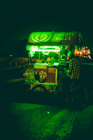 A street-side ice cream cart is illuminated by a bright green light, with a person standing next to it. The cart displays the brand Kwality Wall's and has posters of ice cream products. The background is dark with hints of buildings and people in the distance.
