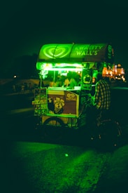 A street-side ice cream cart is illuminated by a bright green light, with a person standing next to it. The cart displays the brand Kwality Wall's and has posters of ice cream products. The background is dark with hints of buildings and people in the distance.