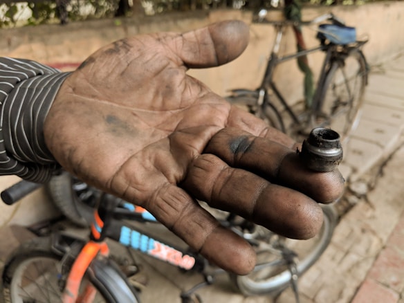 A hand covered in dirt and grease is prominently holding a small metal object, possibly a nut or bolt. In the background, there are bicycles, one of which seems to have parts disassembled. The hand's positioning suggests the person might have been working on repairing or maintaining the bicycles.
