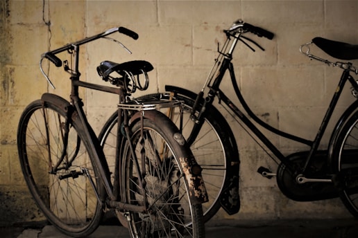 Two vintage bicycles are leaned against a weathered brick wall. The bicycles appear old and rusty, with worn-out seats and tires. The scene has a nostalgic and timeless feel.