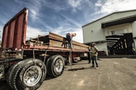 A large flatbed truck is loaded with heavy steel beams outside a warehouse. Three individuals are working around the truck, with one on top of the truck securing the load with chains. The warehouse is labeled with a company name and has an open entrance showing more beams inside. The sky is partly cloudy, casting a dramatic light over the scene.