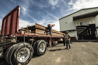 A large flatbed truck is loaded with heavy steel beams outside a warehouse. Three individuals are working around the truck, with one on top of the truck securing the load with chains. The warehouse is labeled with a company name and has an open entrance showing more beams inside. The sky is partly cloudy, casting a dramatic light over the scene.
