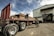 A large flatbed truck is loaded with heavy steel beams outside a warehouse. Three individuals are working around the truck, with one on top of the truck securing the load with chains. The warehouse is labeled with a company name and has an open entrance showing more beams inside. The sky is partly cloudy, casting a dramatic light over the scene.