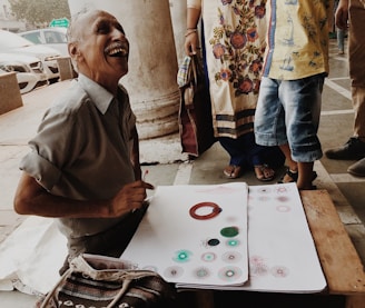 An elderly man sitting on the ground is laughing joyfully while working with a set of drawing tools and papers that have colorful geometric patterns. Beside him, a person wearing a floral-patterned dress is standing with a child wearing denim shorts and a yellow shirt. The background shows part of a car and some architectural columns.