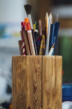 A wooden holder contains various pencils, brushes, and pens of different colors and sizes, standing upright. The surface of the holder features a natural wood grain pattern. The background is softly blurred, emphasizing the holder and its contents.