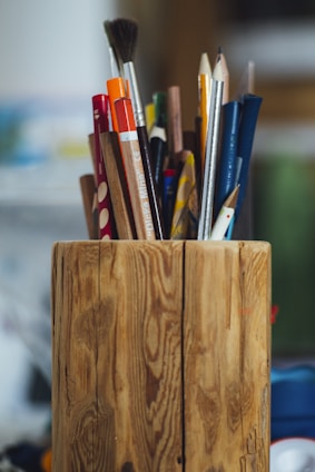A wooden holder contains various pencils, brushes, and pens of different colors and sizes, standing upright. The surface of the holder features a natural wood grain pattern. The background is softly blurred, emphasizing the holder and its contents.