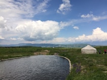 A serene landscape featuring a small body of water surrounded by green grass and hills in the distance. A traditional yurt is positioned on the right side of the image near the water, with two people standing nearby. The sky is vast with large, fluffy clouds scattered across it, adding depth to the scenic view.