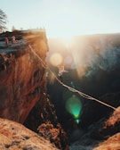 A person balances on a highline stretched between two cliffs, with a dramatic landscape and the warm glow of a setting sun in the background. Onlookers are gathered at the cliff's edge, and the scene is bathed in a golden light.