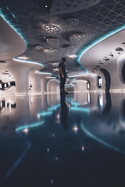 A futuristic hallway with a sleek, modern design featuring intricate ceiling patterns and curved walls. The space is illuminated with ambient blue lighting that reflects off the glossy floor. A solitary person is standing in the center, adding scale to the expansive interior.
