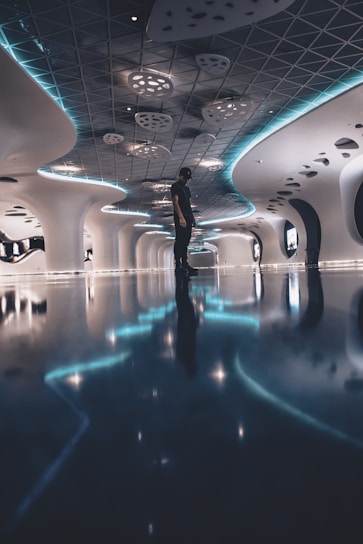 A futuristic hallway with a sleek, modern design featuring intricate ceiling patterns and curved walls. The space is illuminated with ambient blue lighting that reflects off the glossy floor. A solitary person is standing in the center, adding scale to the expansive interior.