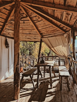 A rustic wooden veranda with a simple table and benches, surrounded by trees with autumn foliage. The space is open and airy, with a curtain gently swaying in the breeze. Sunlight filters through the wooden roof, casting shadows on the floor.