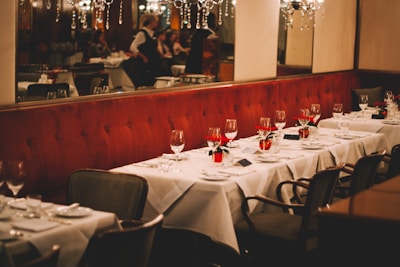 Elegant restaurant interior with tables set for dining, featuring white tablecloths, polished glassware, and red floral centerpieces. Chandeliers hang from the ceiling, and the backdrop has a plush, red cushioned wall.