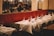 Elegant restaurant interior with tables set for dining, featuring white tablecloths, polished glassware, and red floral centerpieces. Chandeliers hang from the ceiling, and the backdrop has a plush, red cushioned wall.