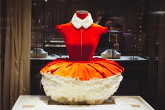 A mannequin dressed in a vibrant red and orange costume with a white frilly skirt is placed inside a display case. The costume has a plush white collar and gold detailing. The background includes a variety of jewelry items on display, with some decorative lights and reflective surfaces, indicating a retail environment.
