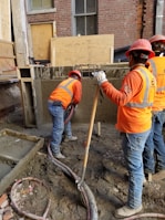 Construction workers wearing orange safety vests and hard hats are working on a building site. One worker is handling a concrete hose while another manages a shovel. The area is filled with wet concrete and building materials.