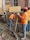 Construction workers wearing orange safety vests and hard hats are working on a building site. One worker is handling a concrete hose while another manages a shovel. The area is filled with wet concrete and building materials.