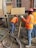 Construction workers wearing orange safety vests and hard hats are working on a building site. One worker is handling a concrete hose while another manages a shovel. The area is filled with wet concrete and building materials.