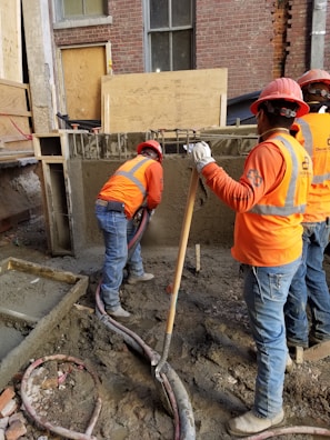 Construction workers wearing orange safety vests and hard hats are working on a building site. One worker is handling a concrete hose while another manages a shovel. The area is filled with wet concrete and building materials.