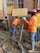 Construction workers wearing orange safety vests and hard hats are working on a building site. One worker is handling a concrete hose while another manages a shovel. The area is filled with wet concrete and building materials.