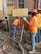Construction workers wearing orange safety vests and hard hats are working on a building site. One worker is handling a concrete hose while another manages a shovel. The area is filled with wet concrete and building materials.