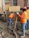 Construction workers wearing orange safety vests and hard hats are working on a building site. One worker is handling a concrete hose while another manages a shovel. The area is filled with wet concrete and building materials.