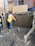 A construction worker wearing a hard hat and safety vest is applying a finish to a freshly laid concrete wall. The area is surrounded by muddy ground, and wooden and metal frameworks are visible. The background includes a brick wall and wooden boards.
