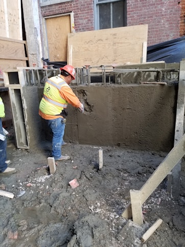 A construction worker wearing a hard hat and safety vest is applying a finish to a freshly laid concrete wall. The area is surrounded by muddy ground, and wooden and metal frameworks are visible. The background includes a brick wall and wooden boards.